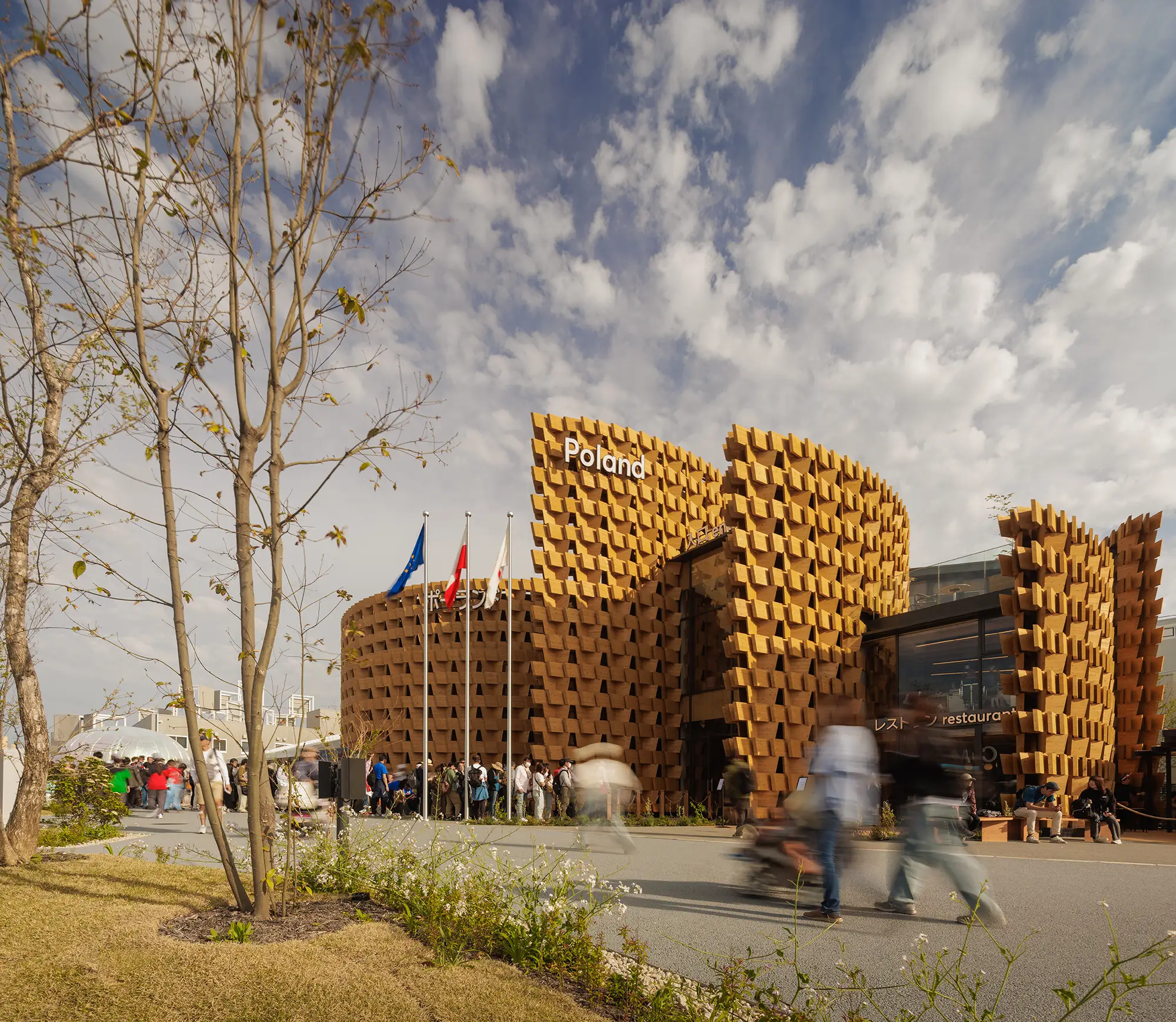 Poland Pavilion viewed from the landscaped Expo grounds, with layered timber walls and moving visitors.