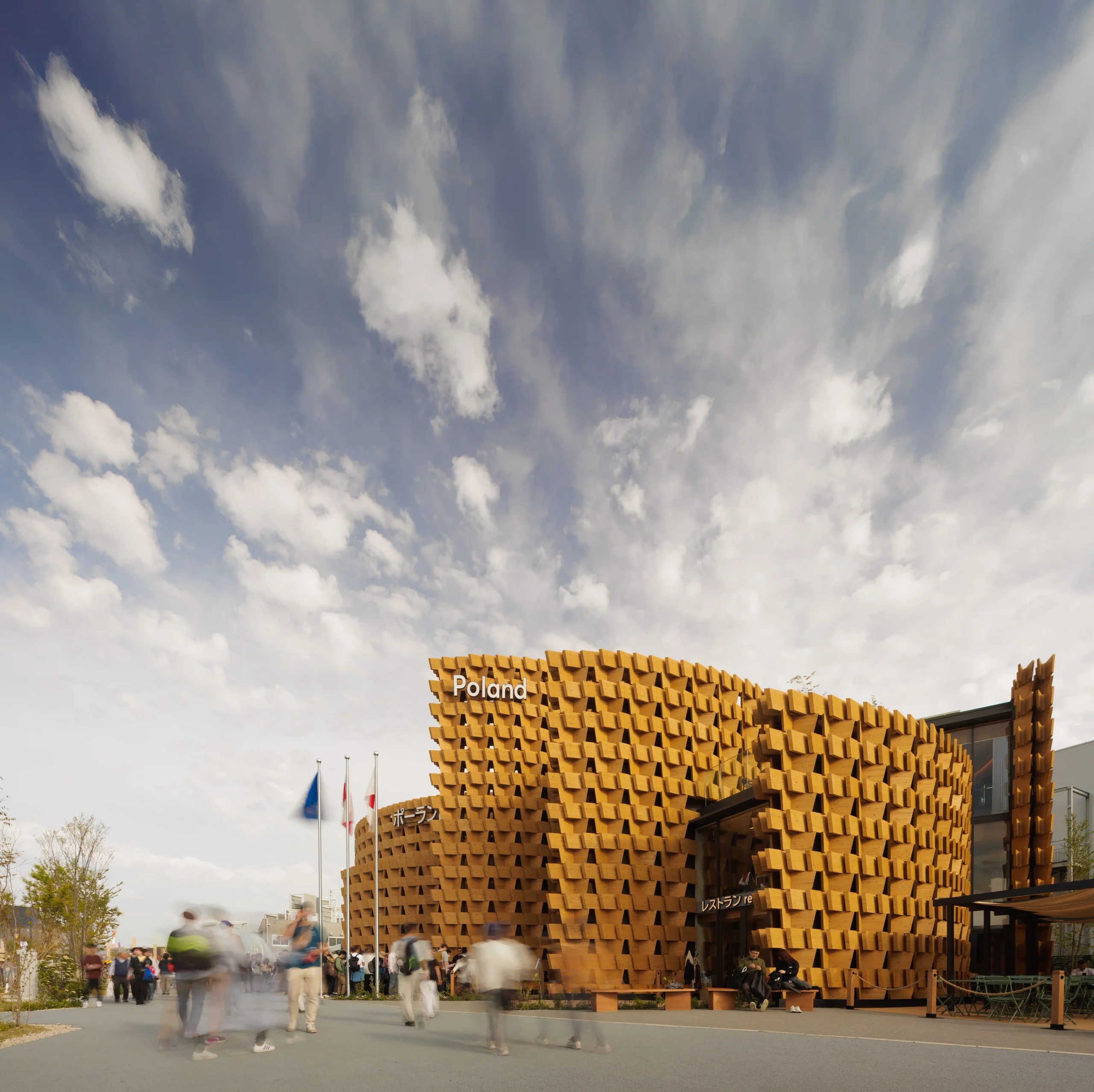Wide exterior view of the Poland Pavilion, showing overlapping curved timber walls beneath a dramatic sky.