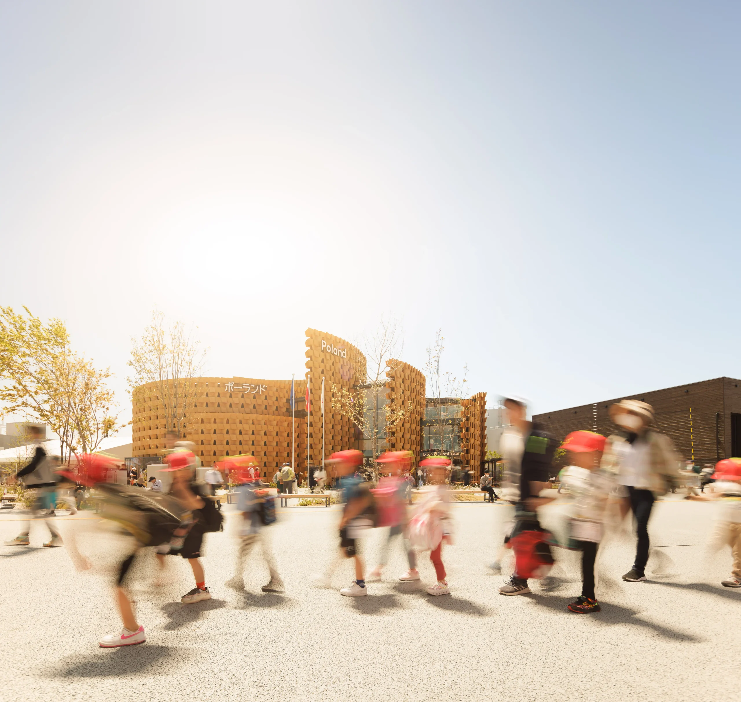 Visitors crossing the foreground in front of the Poland Pavilion, with its timber walls glowing in sunlight.