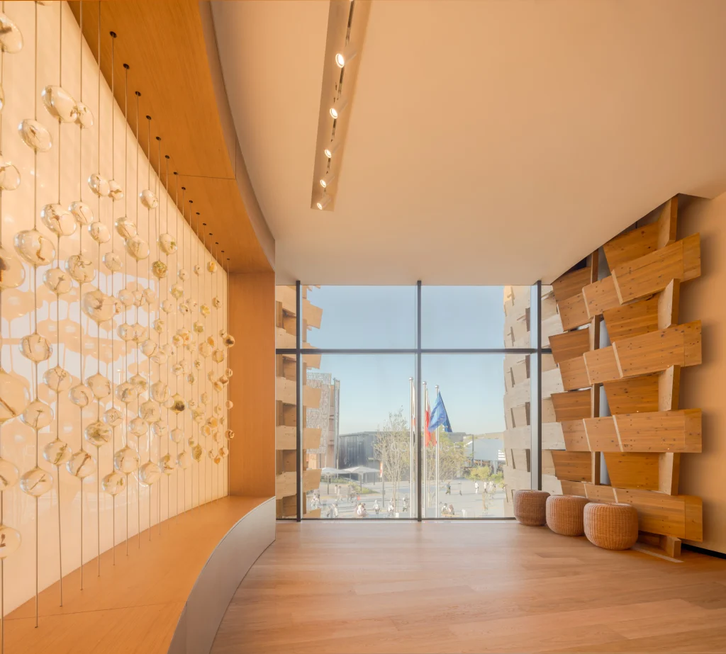 Timber-lined exhibition space with a large window connecting the pavilion interior to the Expo grounds outside.
