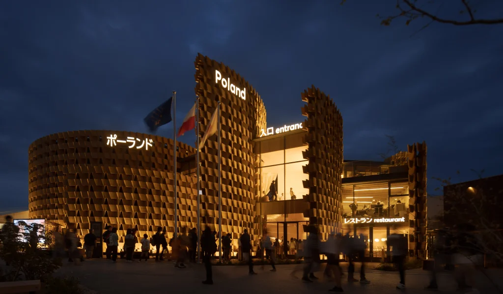 Night view of the completed Poland Pavilion, with illuminated curved timber walls and the Expo entrance area.
