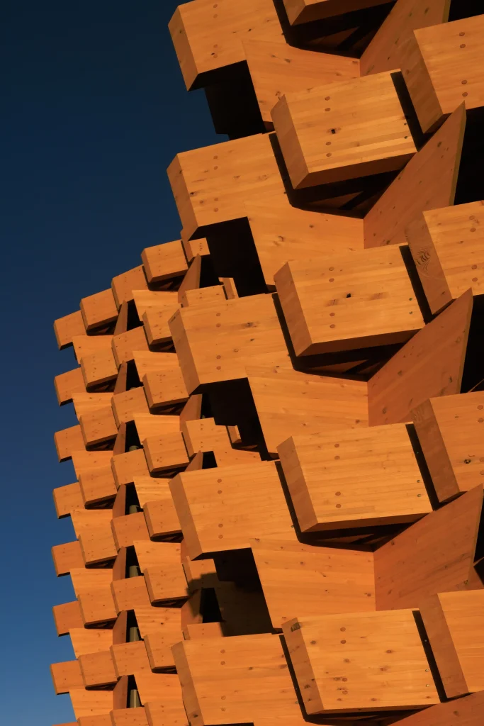 Upward view of the pavilion’s repeating timber façade modules against a clear blue sky.